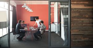 Small team meeting in a modern office boardroom, collaborating on a project around a table with laptops and notes.